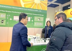 A representative at Zhengzhou Freshliance Electronics Corp., Ltd (1st from the left) is talking with visitors at their booth. / 郑州鲜境电子有限公司的一名代表（左一）正在展位上与参观者交谈。